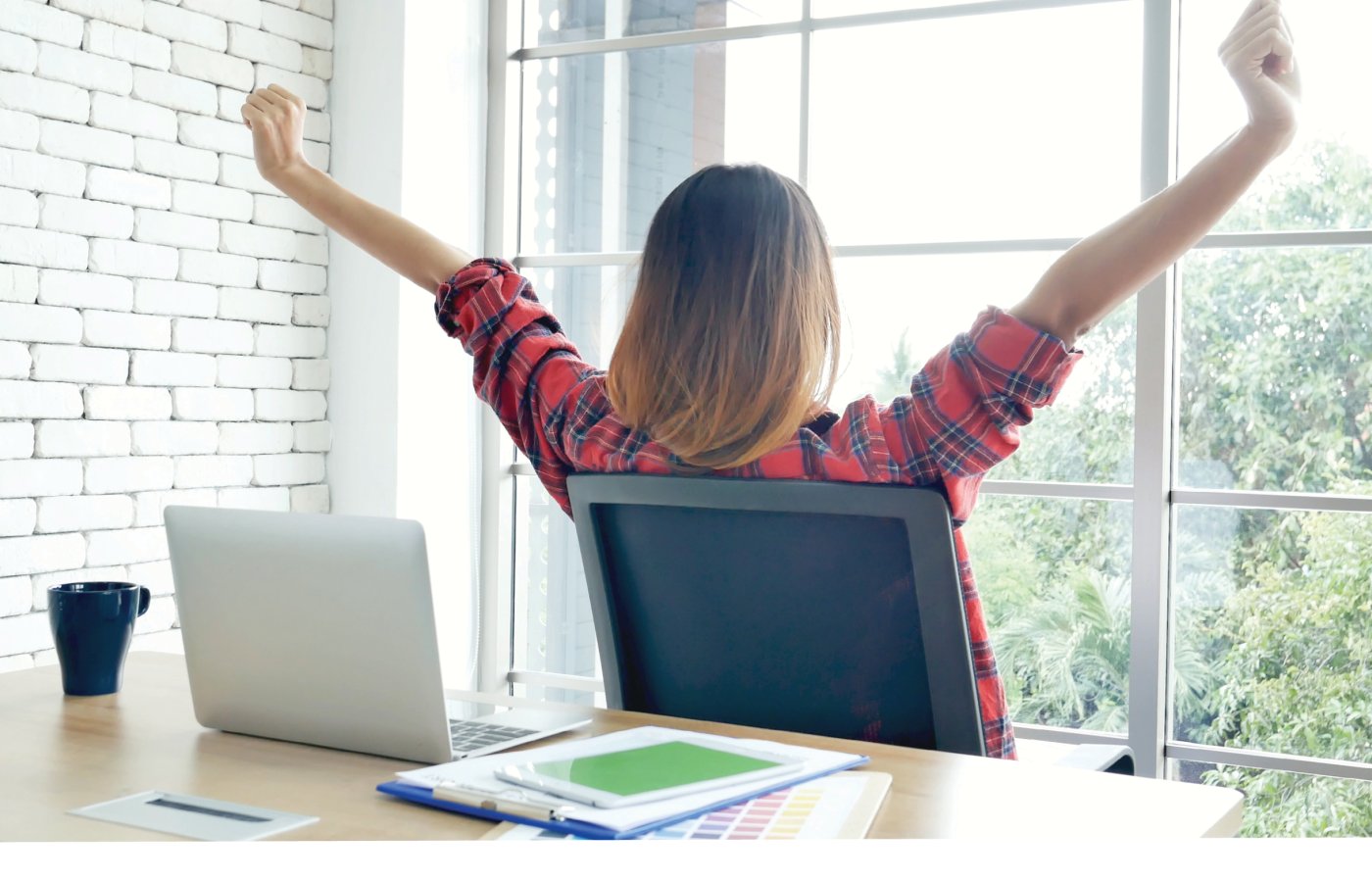 woman at desk looking out window with arms raised in the air.
