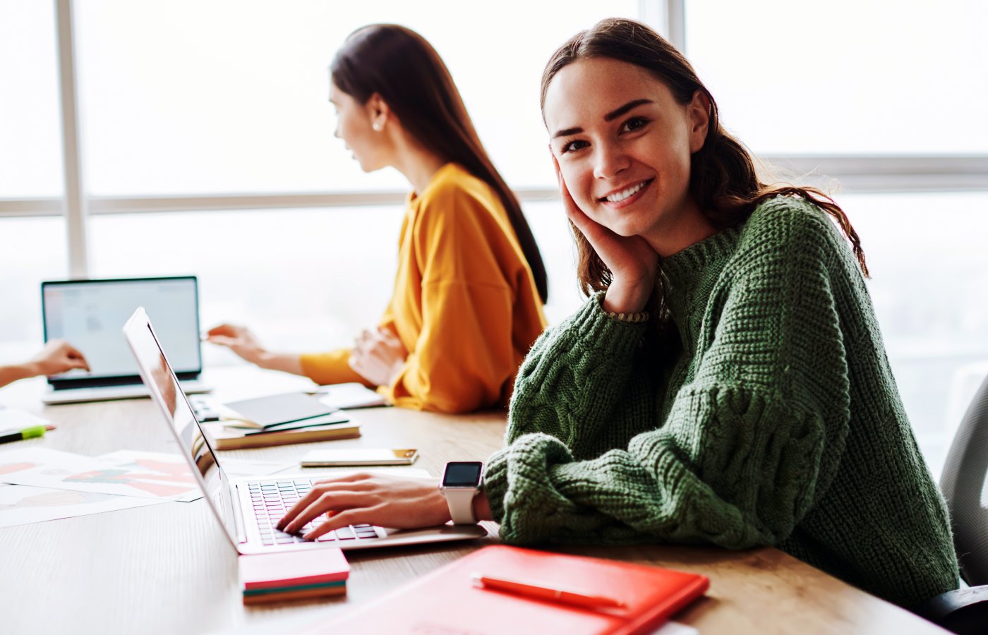 Young woman working at a laptop with other people nearby and is smiling at the camera