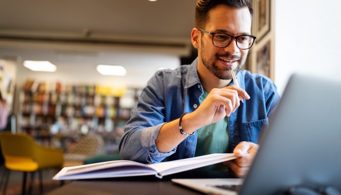 young man in a library looking at a laptop with open books