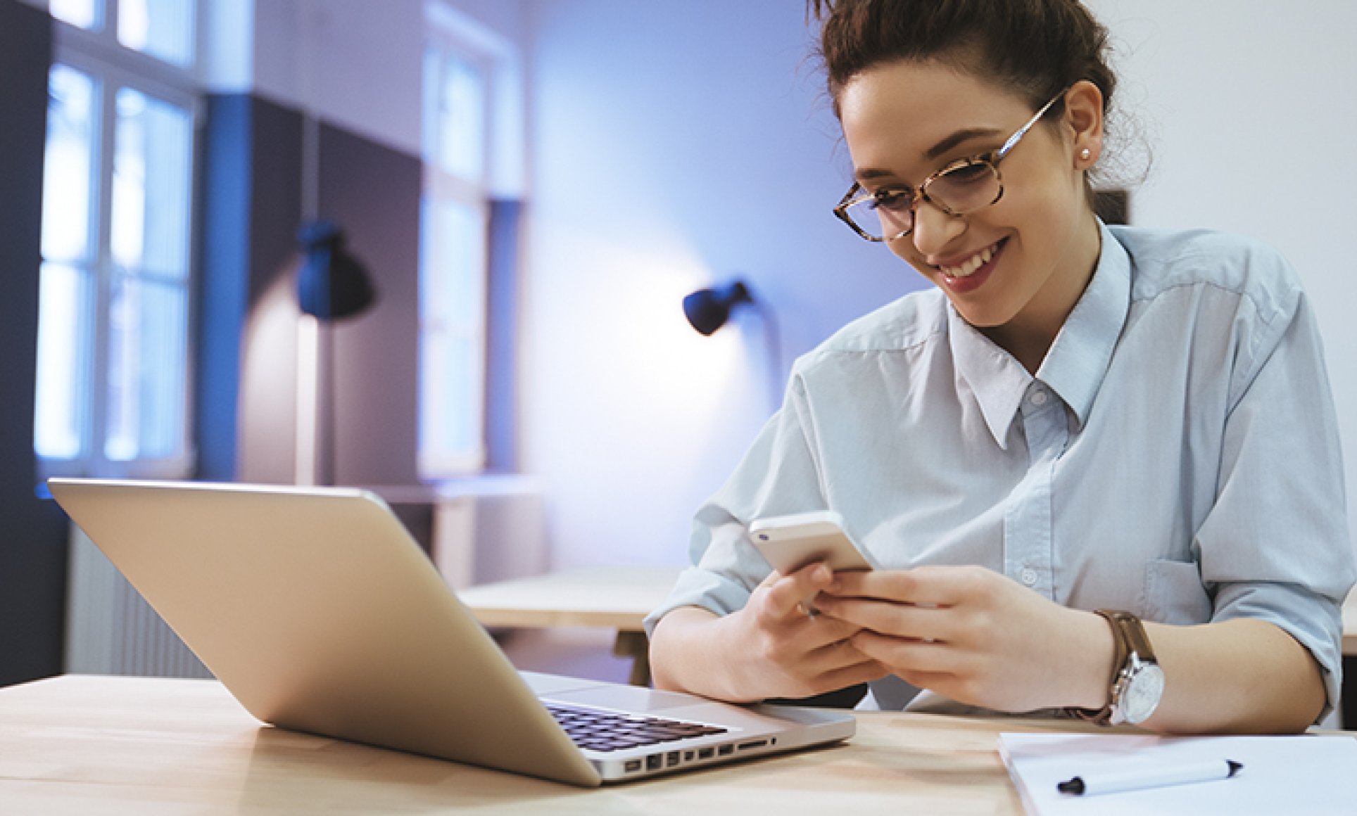 woman smiling, looking at phone in front of laptop