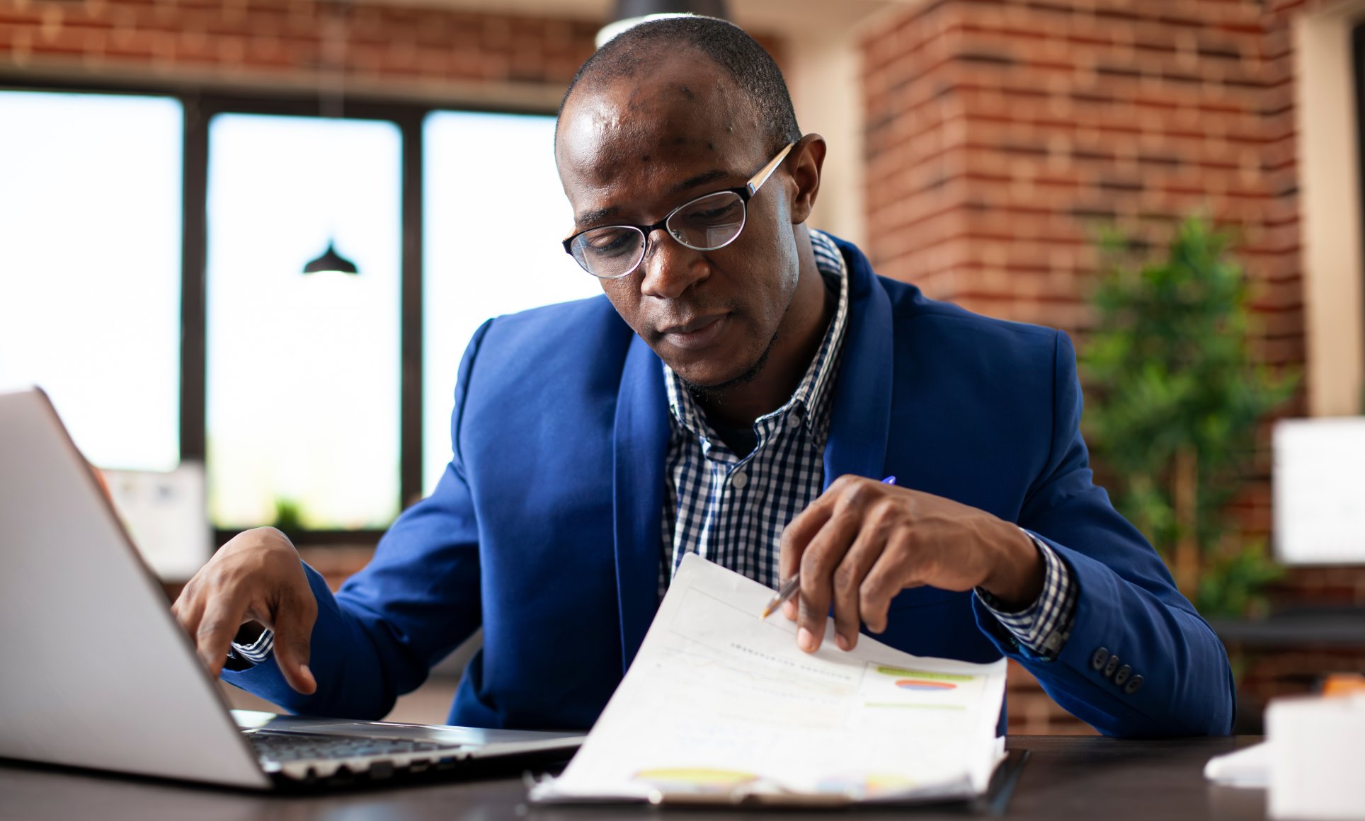 Man closely looking through papers while sitting at a desk with a laptop. 