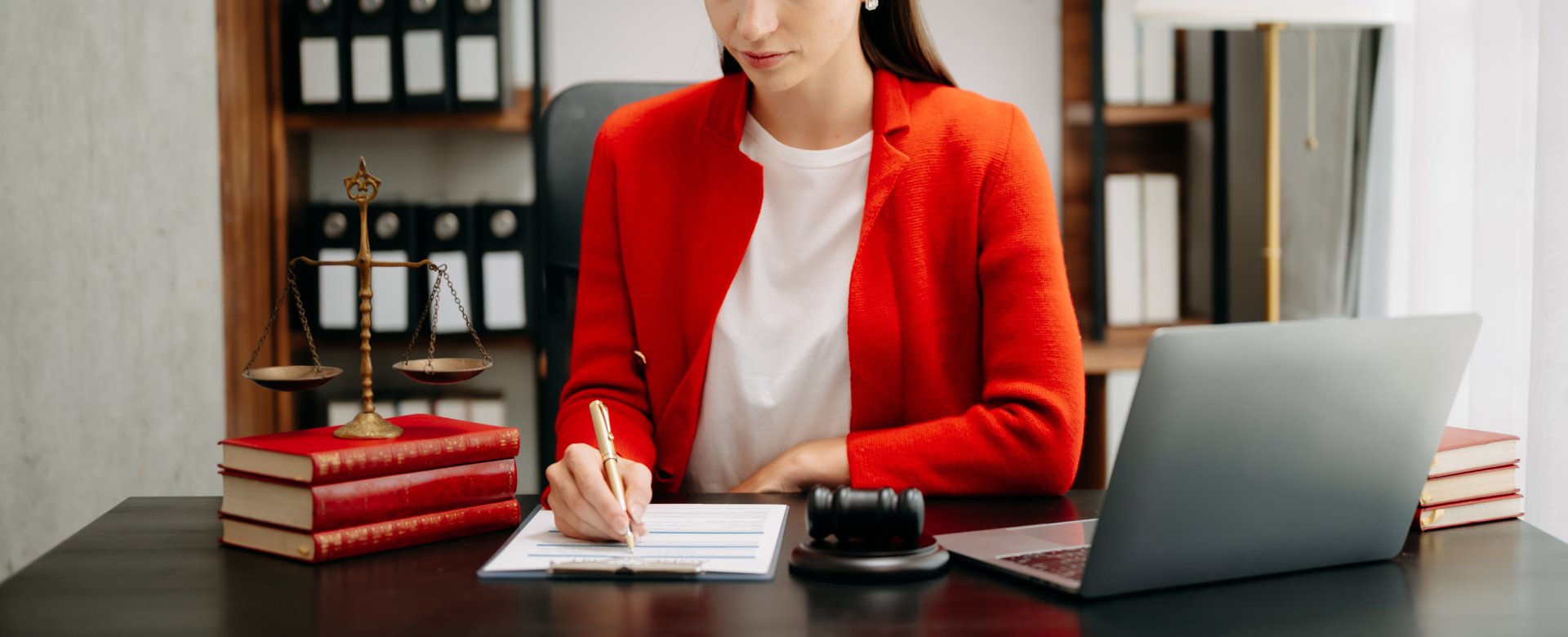woman writing on a piece of paper while seated at a desk. A laptop, gavel, law books, and scale are placed on the desk.  