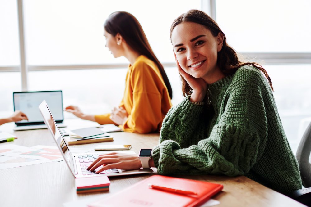 Young woman working at a laptop with other people nearby and is smiling at the camera